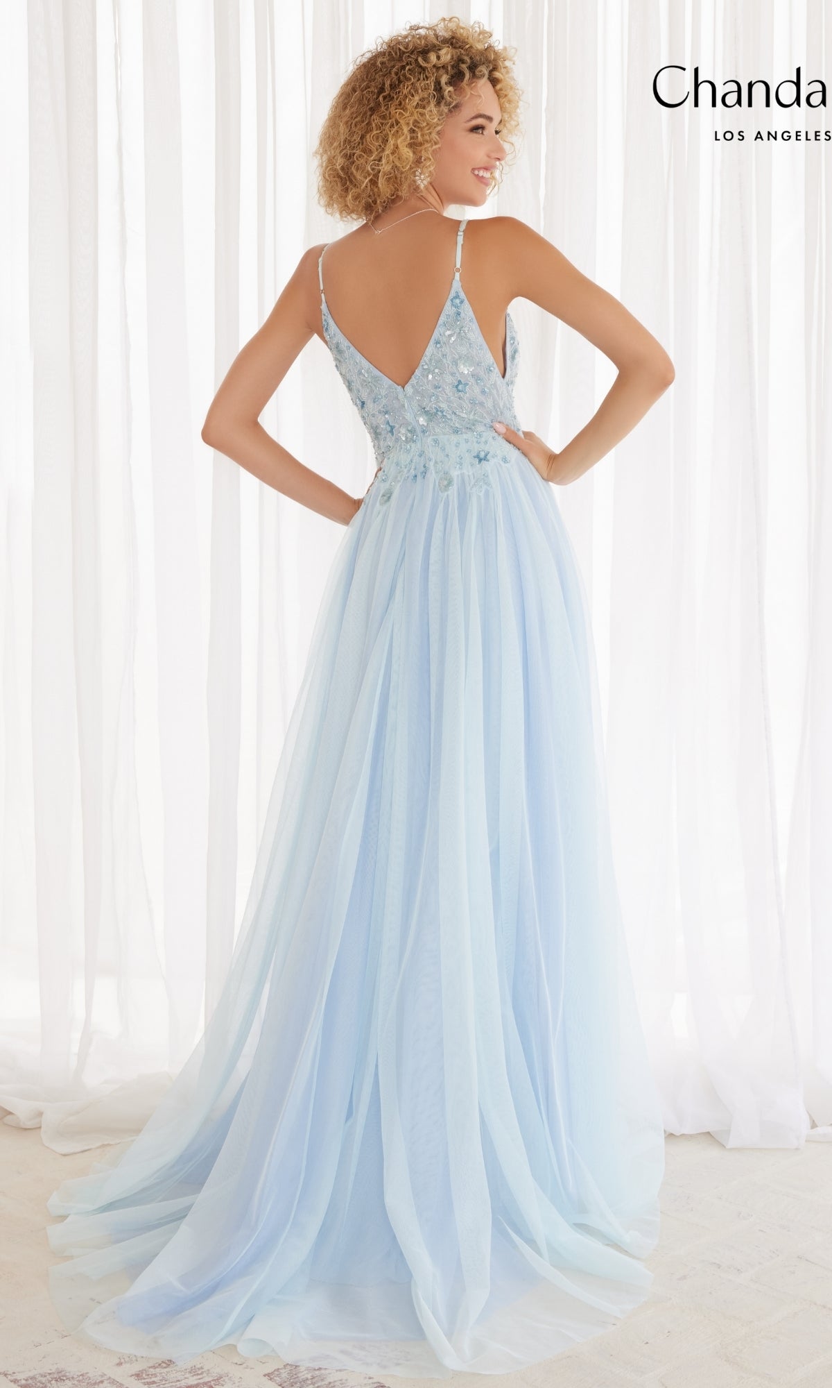 A woman stands before sheer white curtains wearing the Long Prom Dress 30165 by Chandalier—a light blue A-line formal gown featuring a deep V-back and delicate floral embellishments.